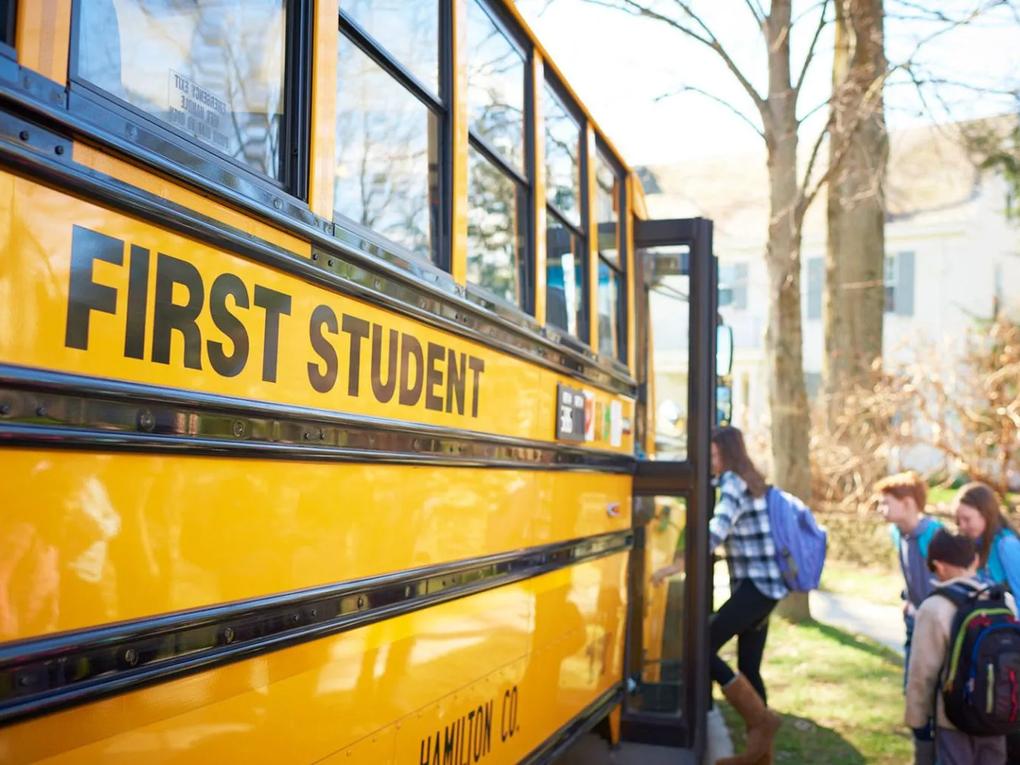 Children boarding a yellow school bus labeled "First Student" on a sunny day.