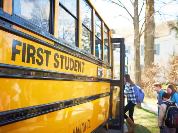Children boarding a yellow school bus labeled "First Student" on a sunny day.