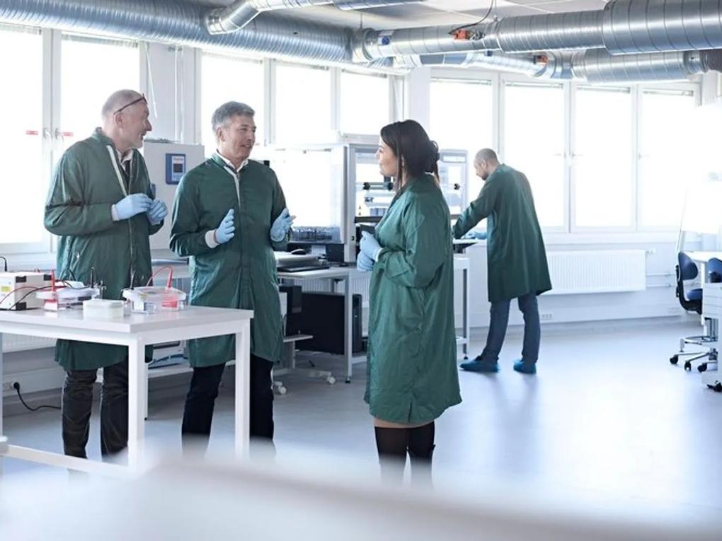 Three scientists in green lab coats and gloves are having a discussion in a bright laboratory.