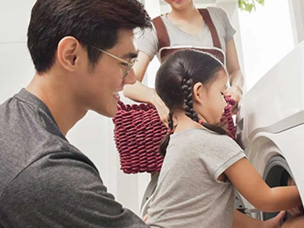 A man smiling as he helps a young girl load laundry into a washing machine.