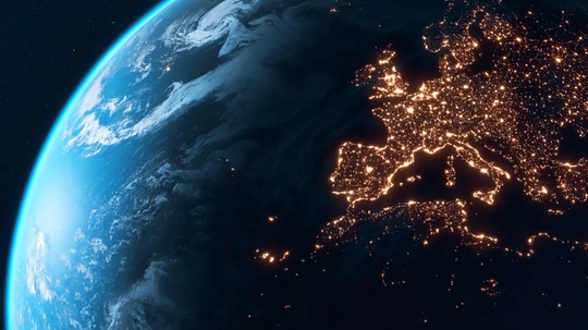 A view of Earth from space at night, showing illuminated cities across Europe with a visible cloud cover over the Atlantic Ocean.