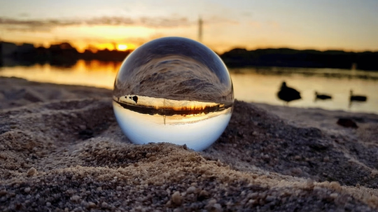 A clear glass sphere on sandy ground reflecting a sunset over a lake, with silhouettes of ducks in the background.