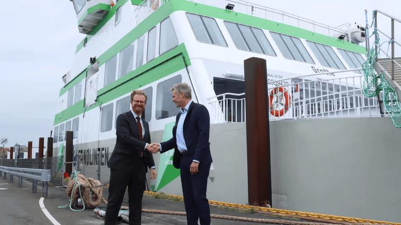 Two men in suits shaking hands in front of a large green and white ferry docked at a port.