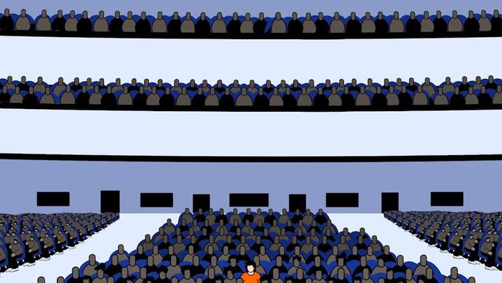 A figure in an orange shirt sits among many figures wearing dark clothes.