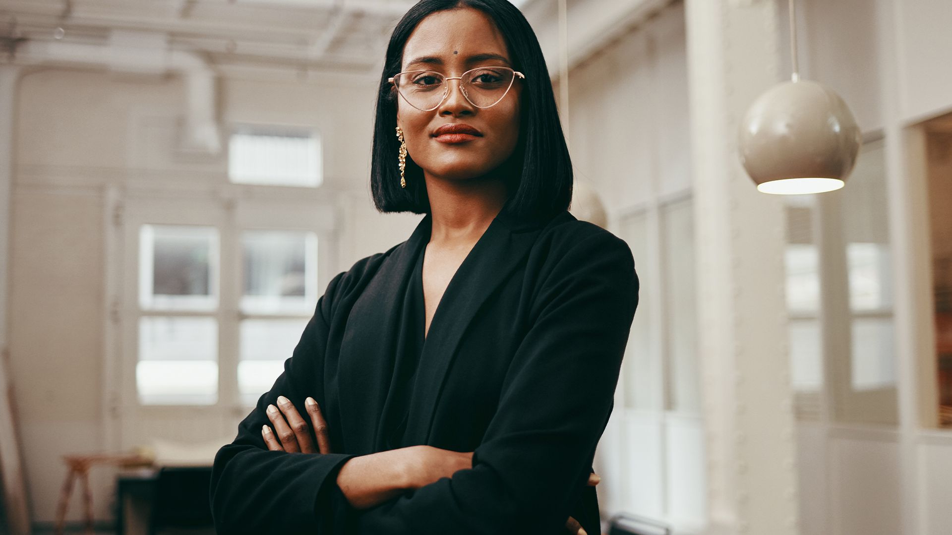 A person with glasses and short hair stands with arms crossed in a bright office space.
