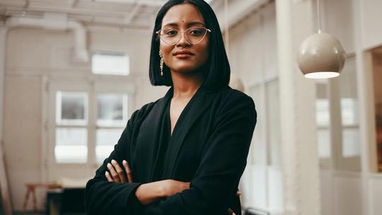 A person with glasses and short hair stands with arms crossed in a bright office space.