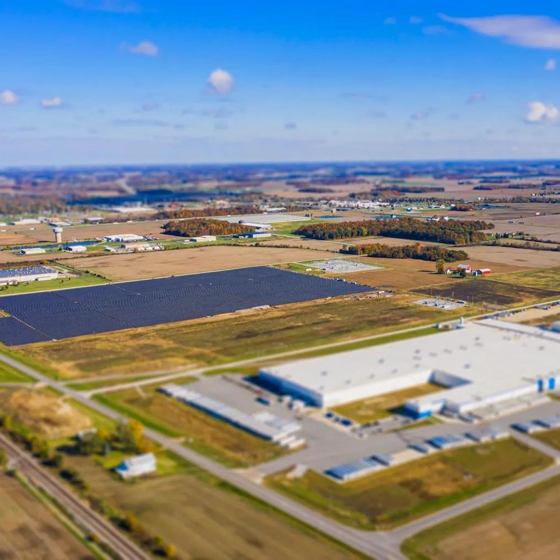 Aerial view of a large industrial complex surrounded by fields and roads, with a solar panel array visible in the center.