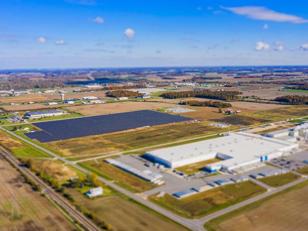 Aerial view of a large industrial complex surrounded by fields and roads, with a solar panel array visible in the center.