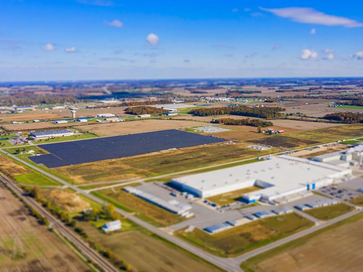 Aerial view of a large industrial complex surrounded by fields and roads, with a solar panel array visible in the center.