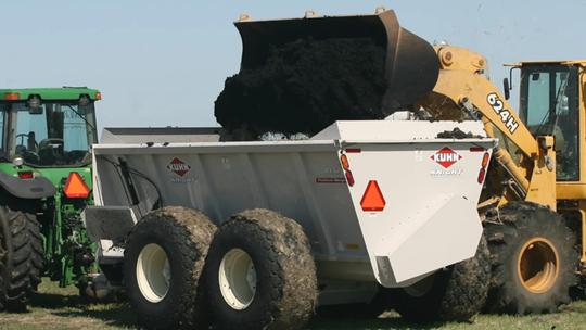 A tractor with a large white trailer being loaded with dark soil by a yellow loader on a grassy field.