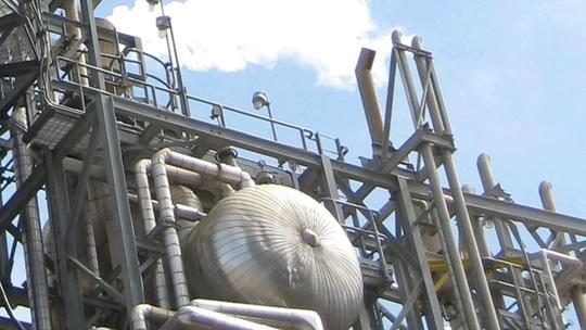 Industrial structure with metal pipes and tanks against a blue sky, emitting steam or smoke, showcasing a section of a processing plant or refinery.