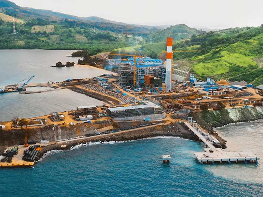 Aerial view of a coastal industrial construction site with cranes, scaffolding, and a tall chimney.