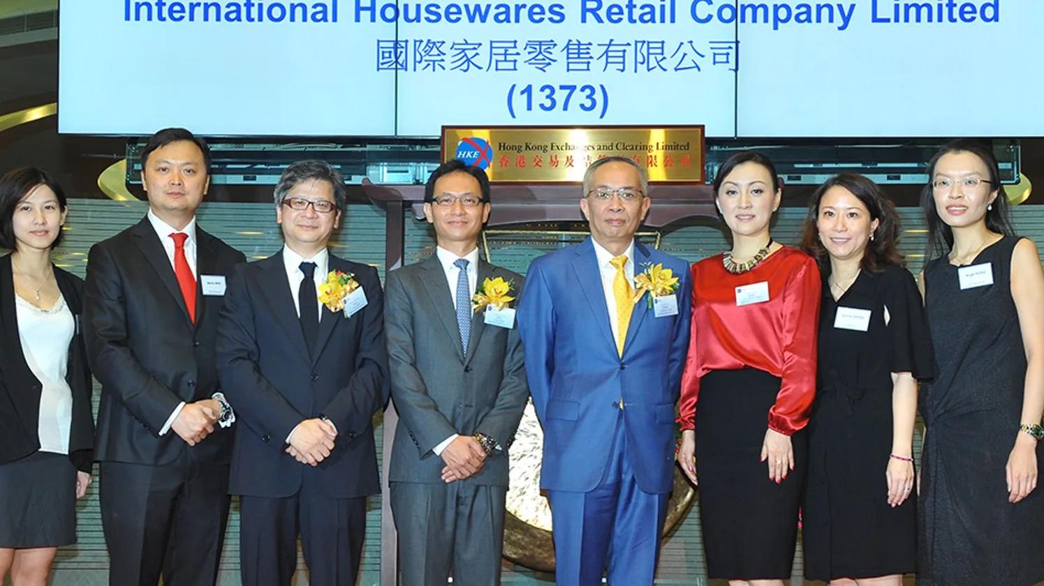 A group of executives and professionals standing together at an event, celebrating the listing of International Housewares Retail Company Limited, with a sign in the background displaying the company's name and stock code.