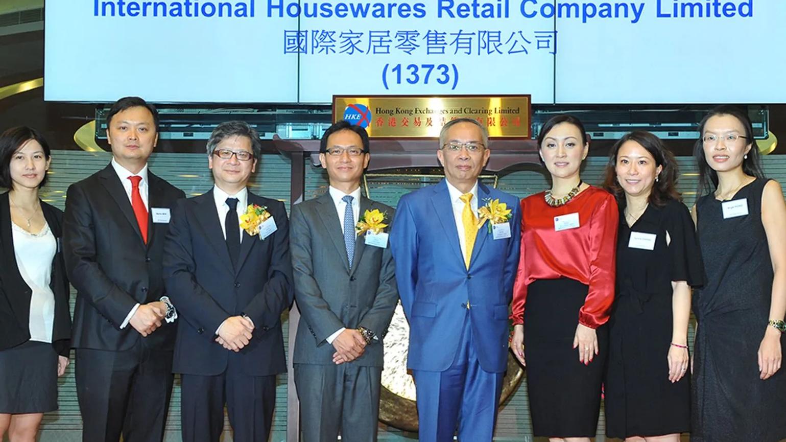 A group of executives and professionals standing together at an event, celebrating the listing of International Housewares Retail Company Limited, with a sign in the background displaying the company's name and stock code.