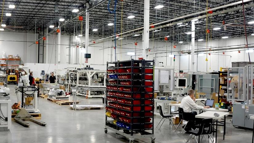 A spacious industrial warehouse with machinery, shelves of parts, and a person working at a desk with a laptop.