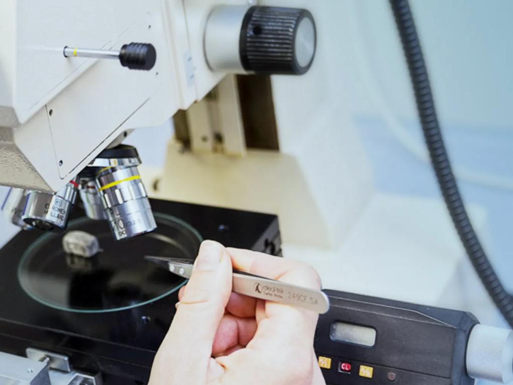 A person using tweezers to handle a sample under a microscope in a laboratory setting.