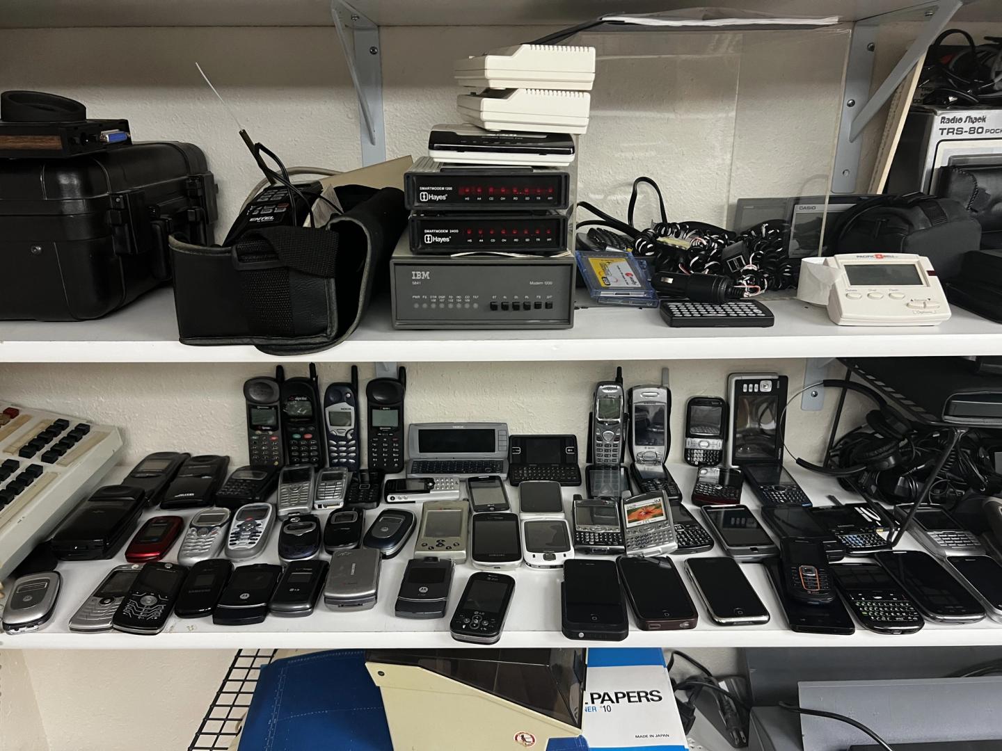 two shelves of obsolete telecommunication like an IBM modem and various PDAs and early cell phones