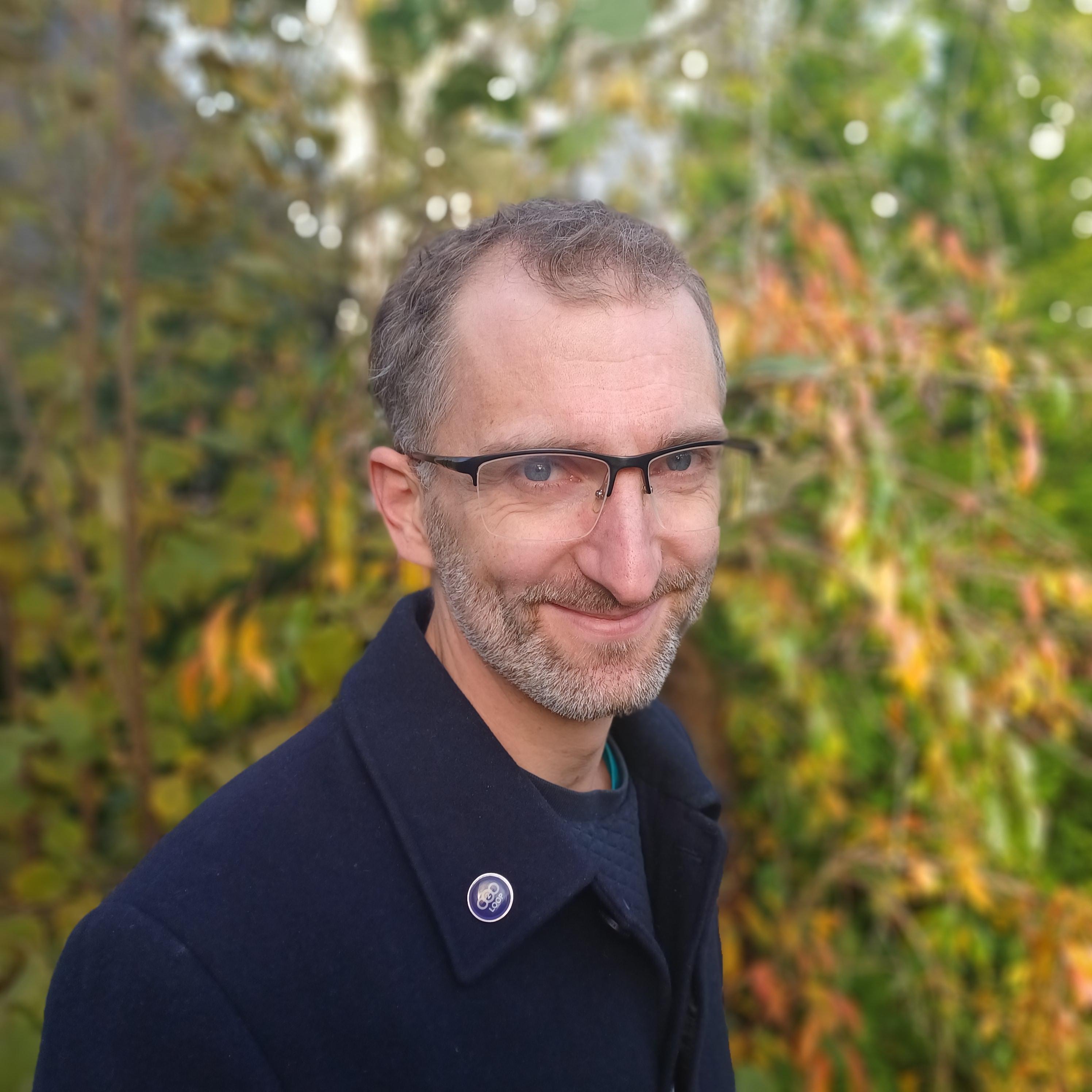 Smiling man with glasses and a beard wearing a dark coat, against blurred autumn foliage.