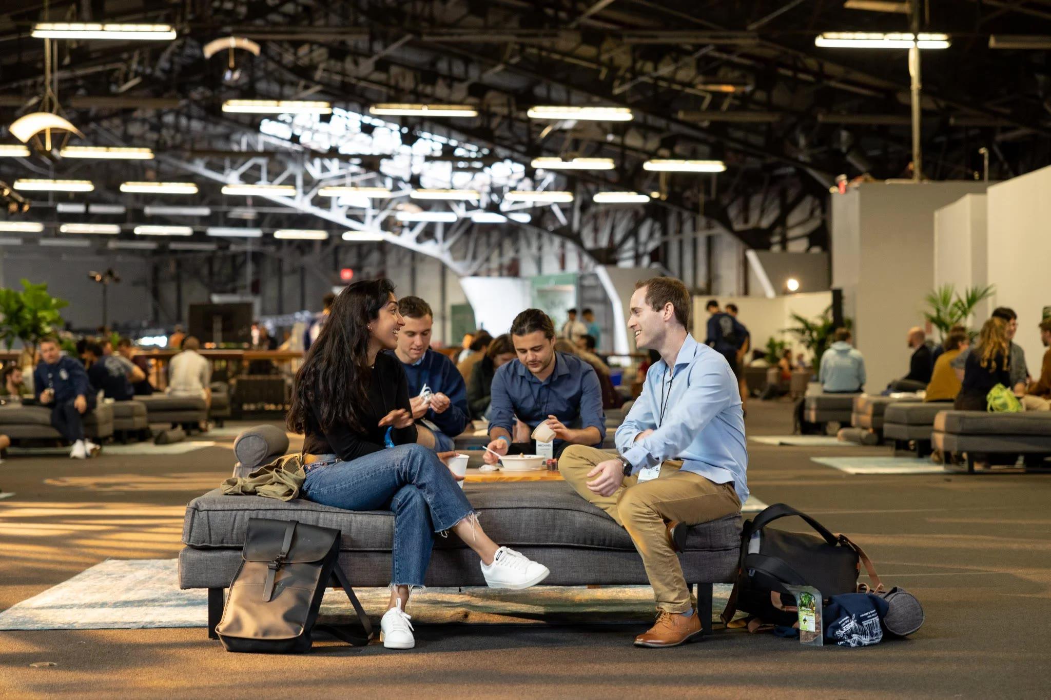 Een foto waarbij centraal twee mensen een gesprek hebben in een soort event hall met andere mensen op de achtergrond.