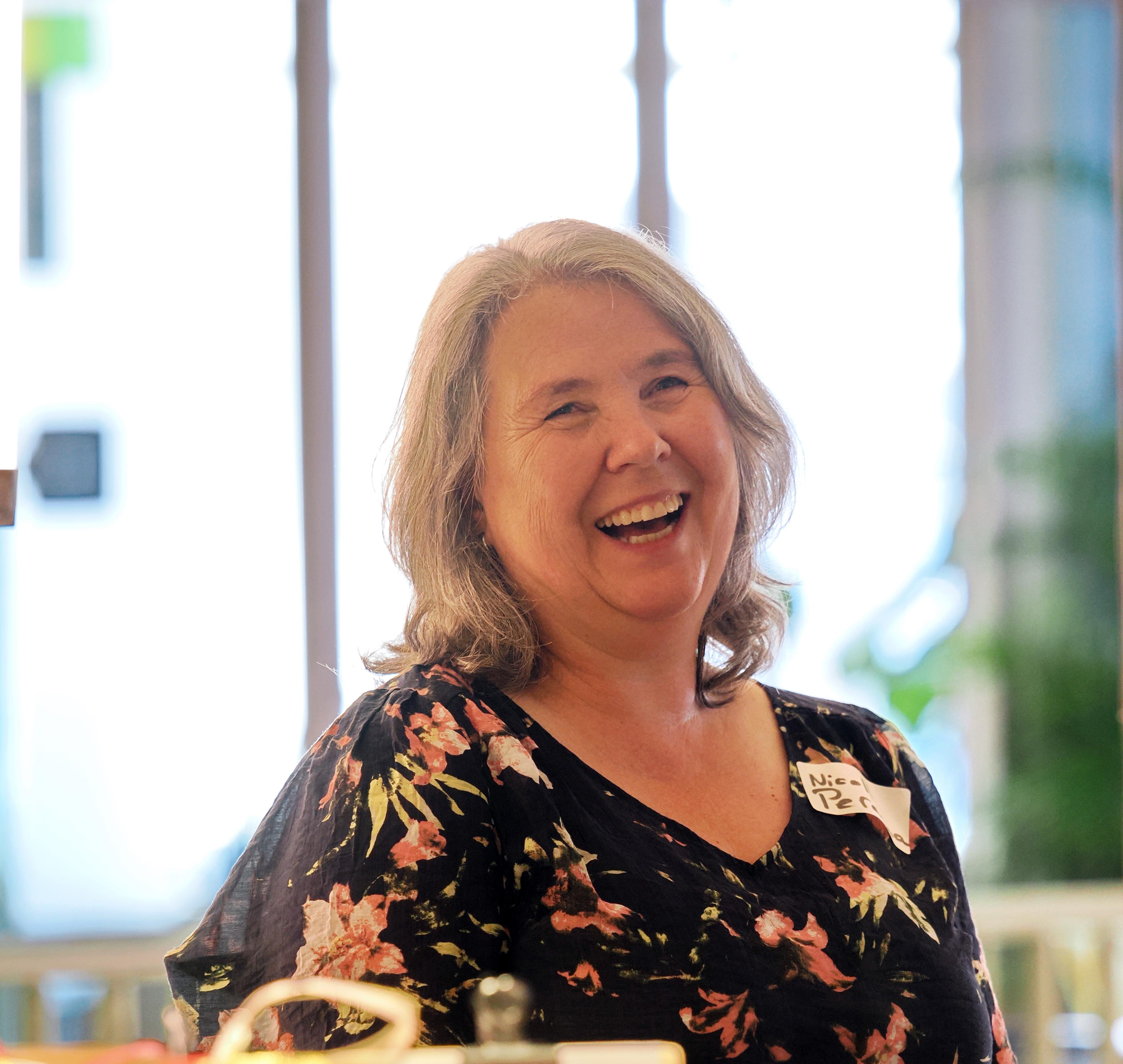 A woman with grey hair and a floral top laughs brightly, standing behind a table.