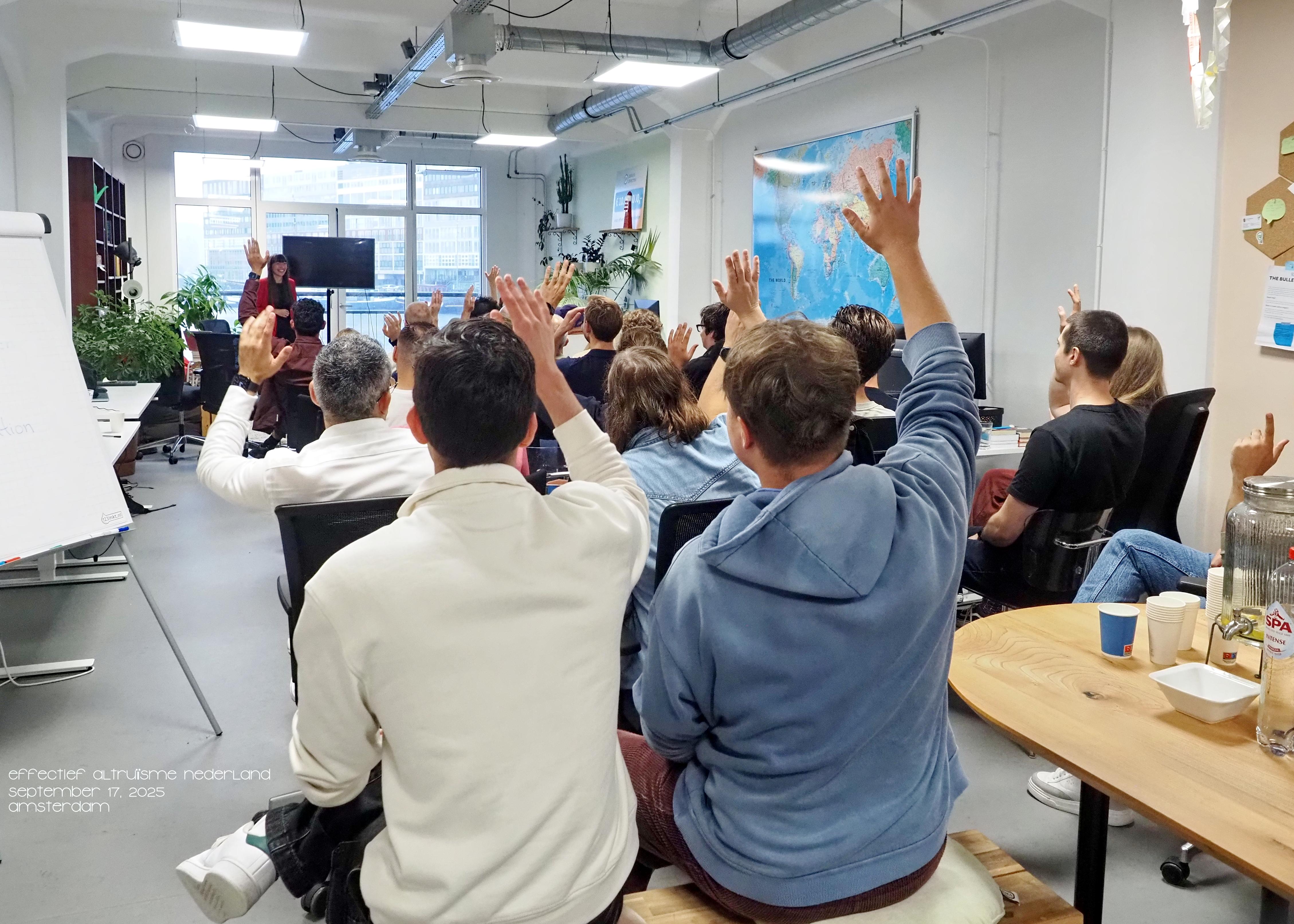 A group of people in a bright room with many hands raised, facing a presenter.