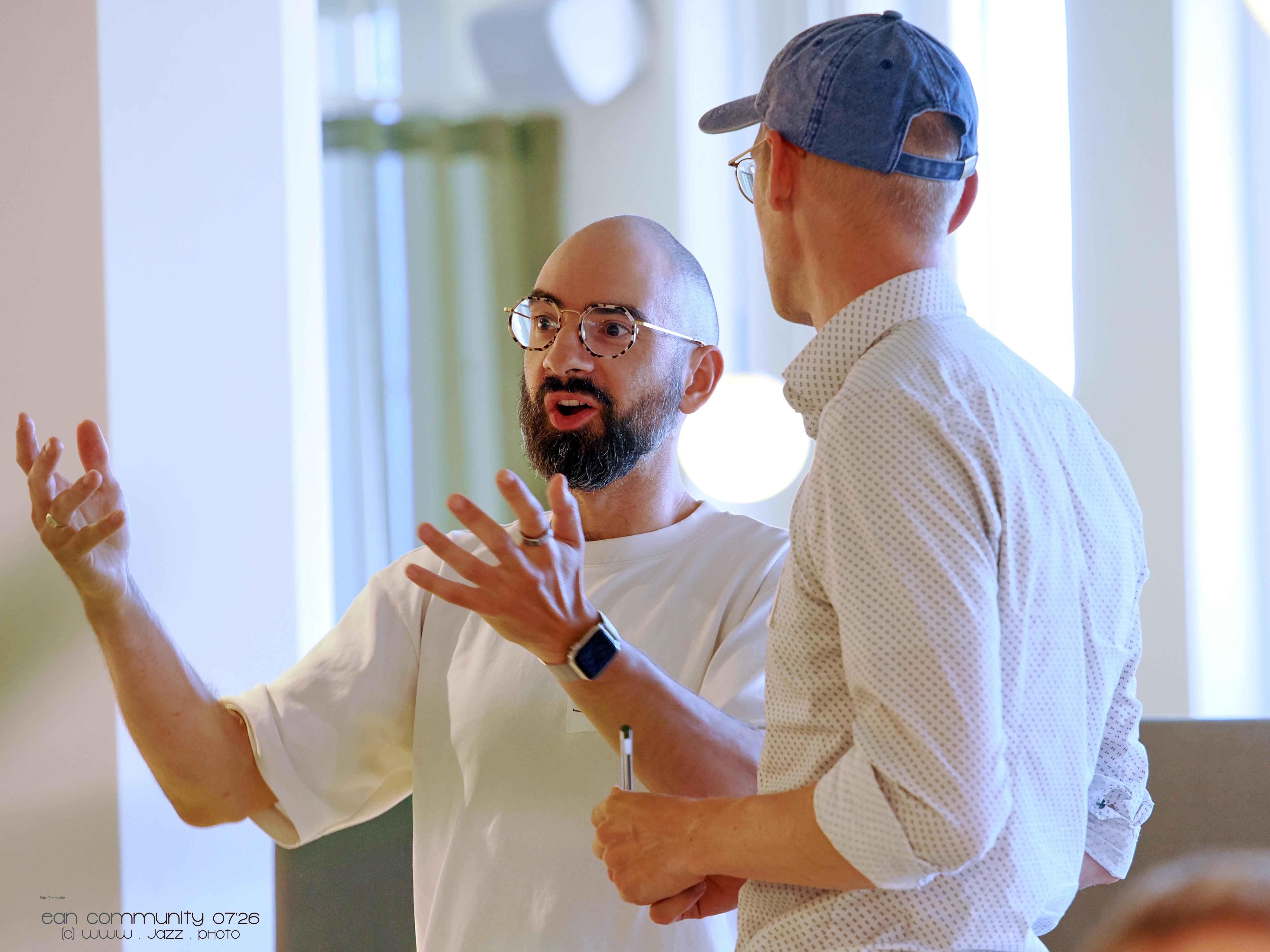 A man with a beard and glasses gestures animatedly while speaking to another man in a baseball cap, seen from behind.