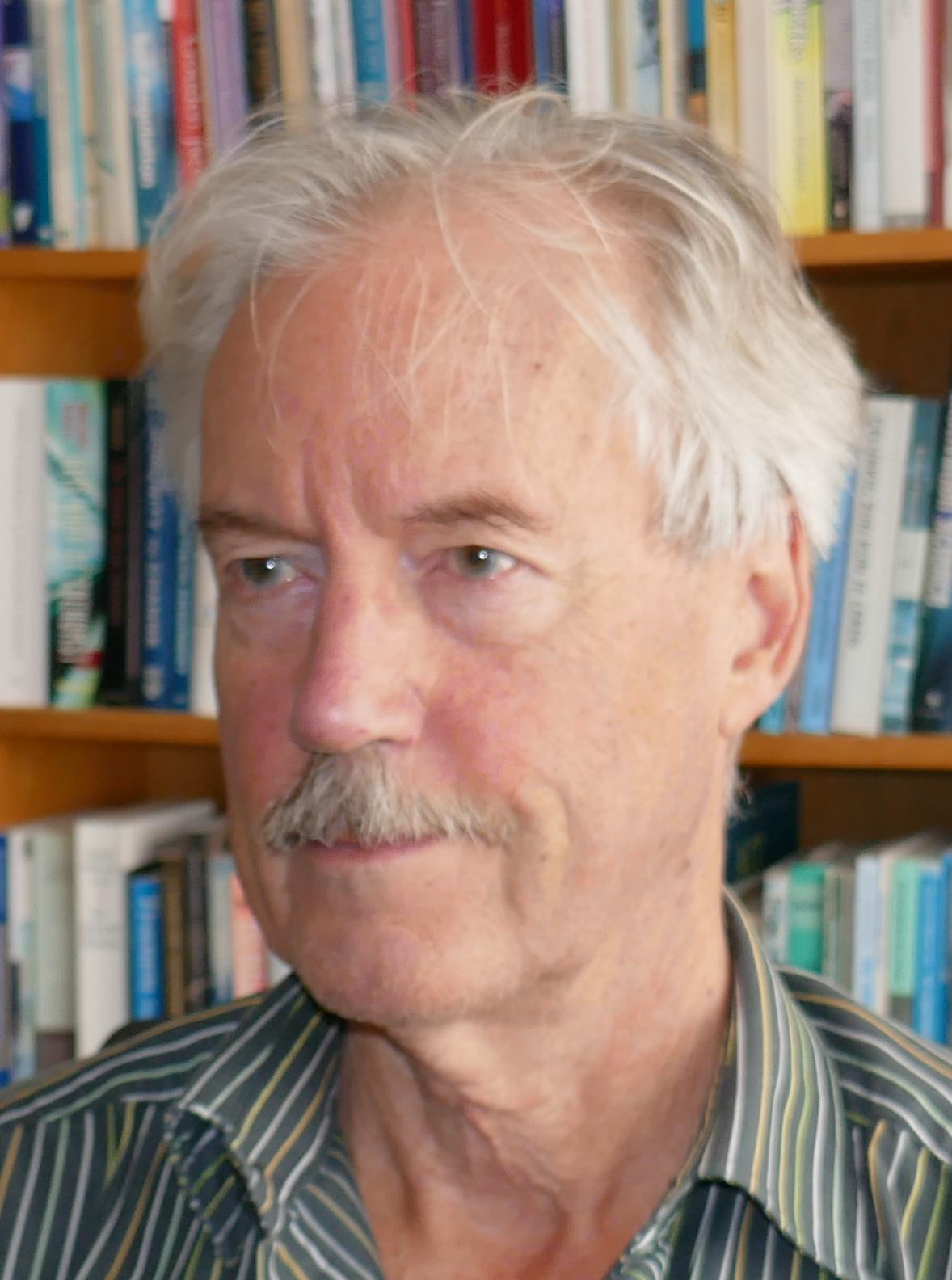 a man with a mustache is standing in front of a bookshelf