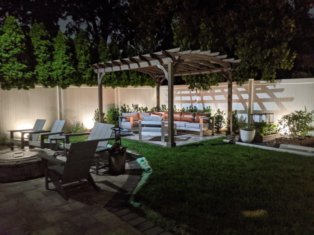 A pergola in the corner of a backyard with exterior landscape uplights along a white fence