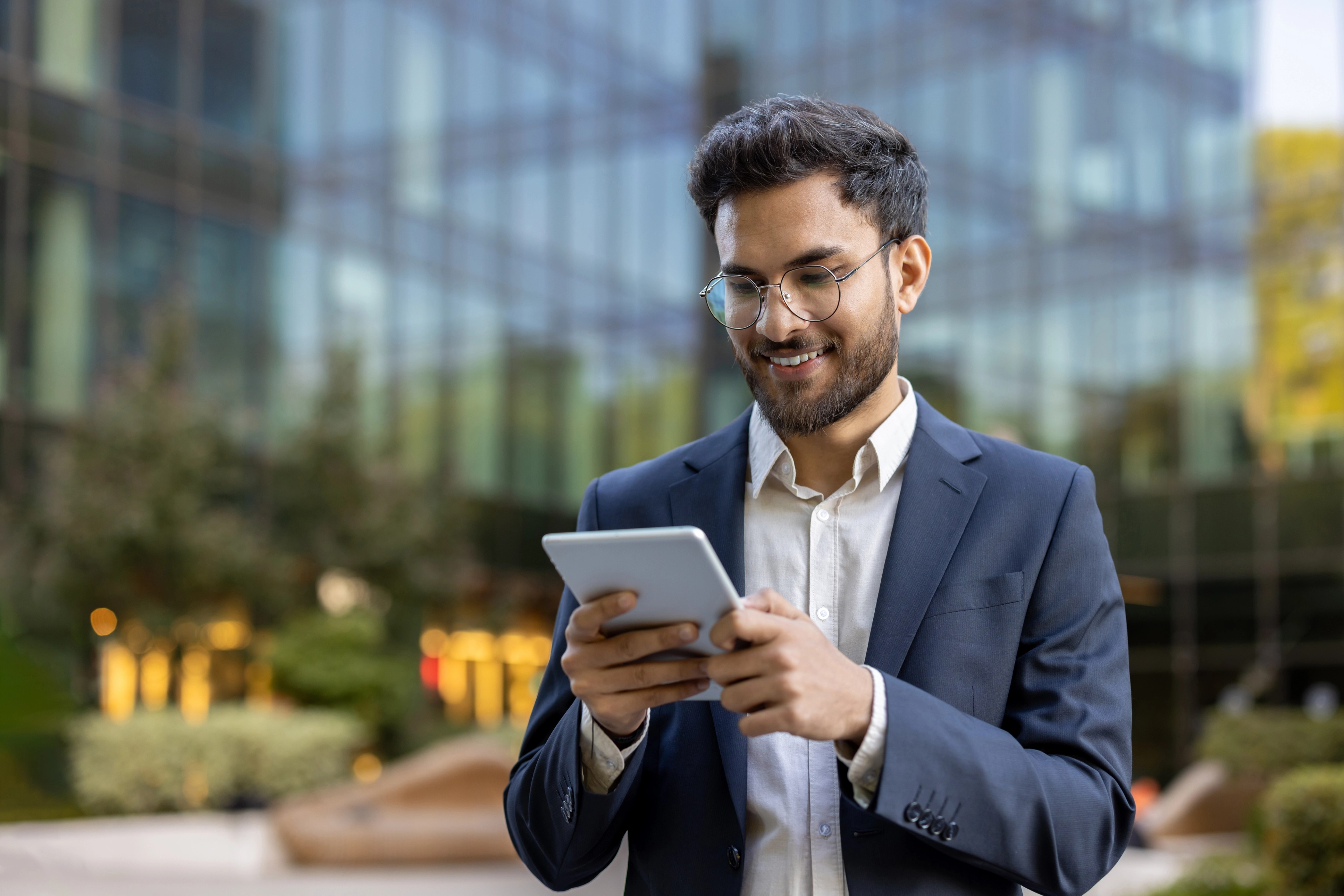 Smiling businessman using digital tablet outside modern glass office building.