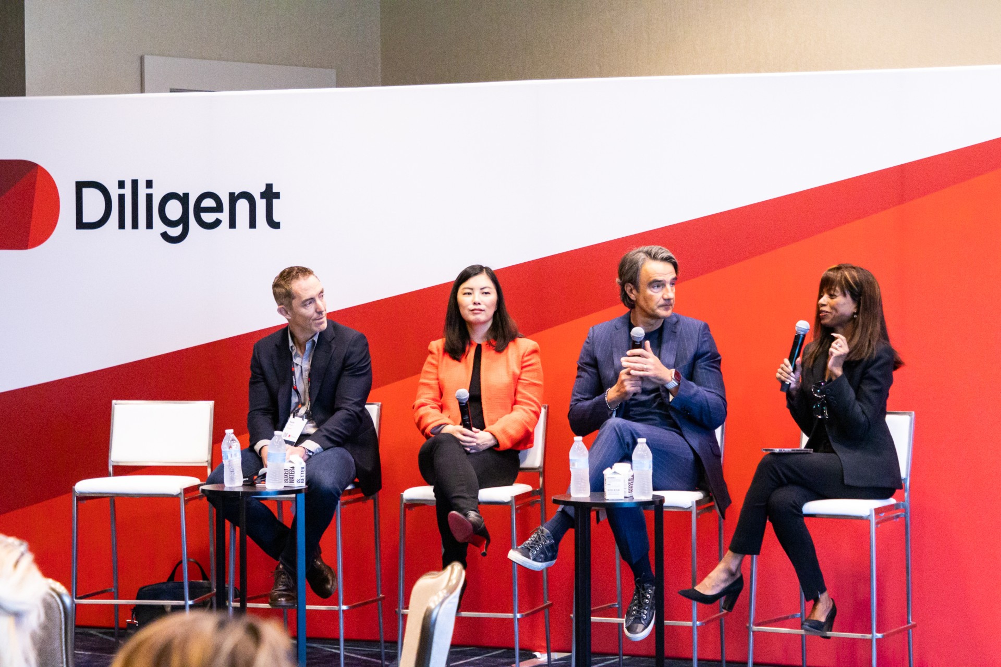 Panelists sit on stools upon a stage holding microphones and addressing an audience