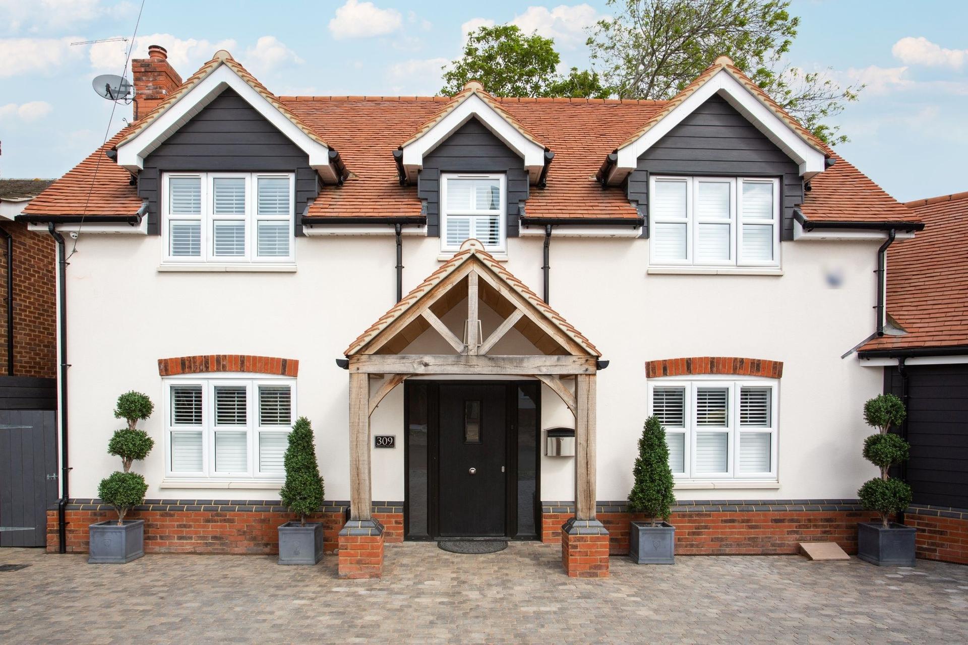 A detached cream-rendered house with a red clay tiled roof, dark-clad dormers and an oak-framed porch.