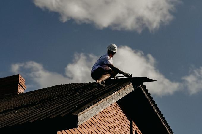 A roofer in a white hard hat works along the edge of a pitched roof on a brick house against a blue sky.