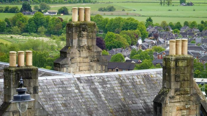 Stone chimney stacks with clay pots on a slate roof overlooking a town and green fields.