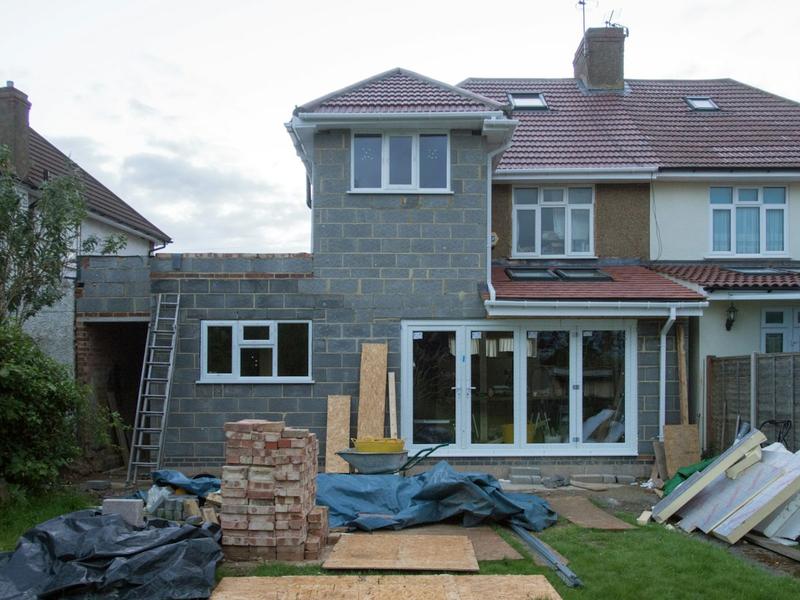 A blockwork side and rear extension under construction on a semi-detached house, with building materials in the garden.