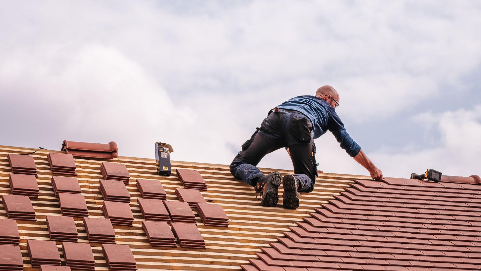 A roofer kneels on roof battens laying red clay plain tiles on a pitched roof under a cloudy sky.