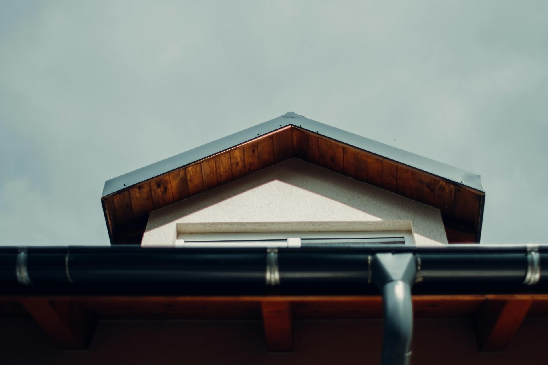 Looking up at a gable with timber soffits and a black gutter and grey downpipe against an overcast sky.