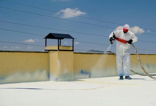 A worker in a white protective suit spray-coats a flat roof with a waterproof finish.