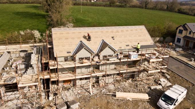 An aerial view of a house under construction wrapped in scaffolding, with roofers working on the battened roof.