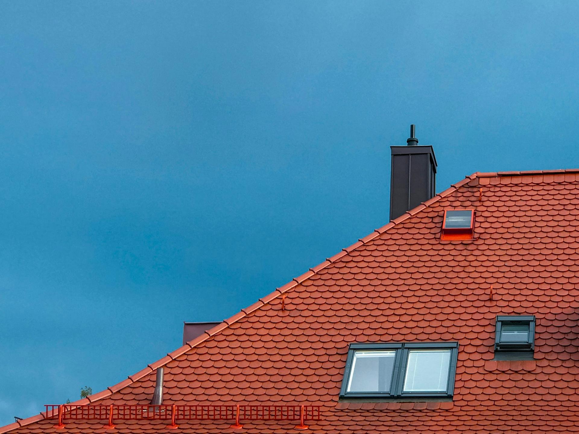 A red clay tiled roof with roof windows and a dark chimney against a deep blue sky.