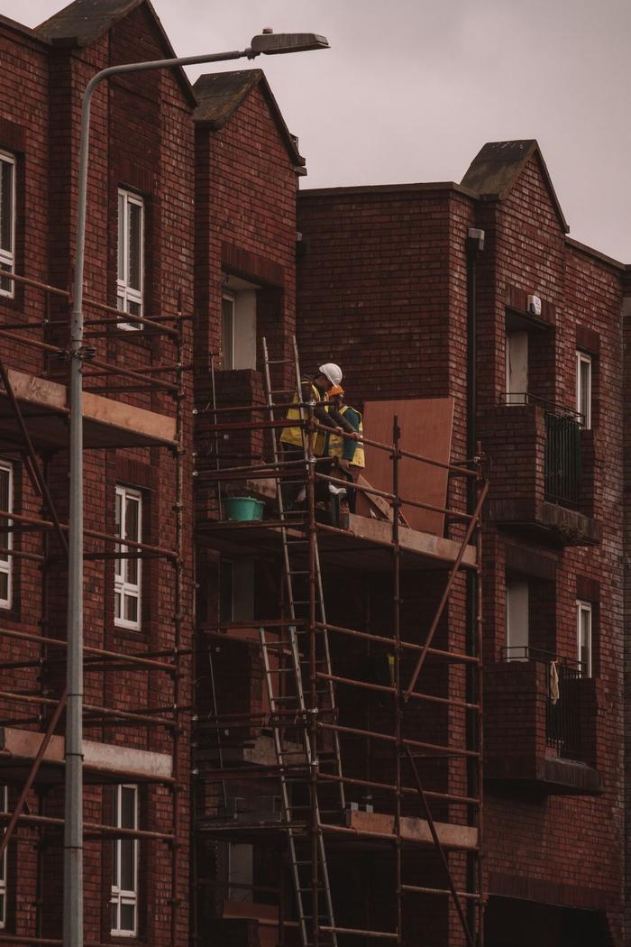Two builders in hi-vis vests work on scaffolding in front of a row of red brick houses.