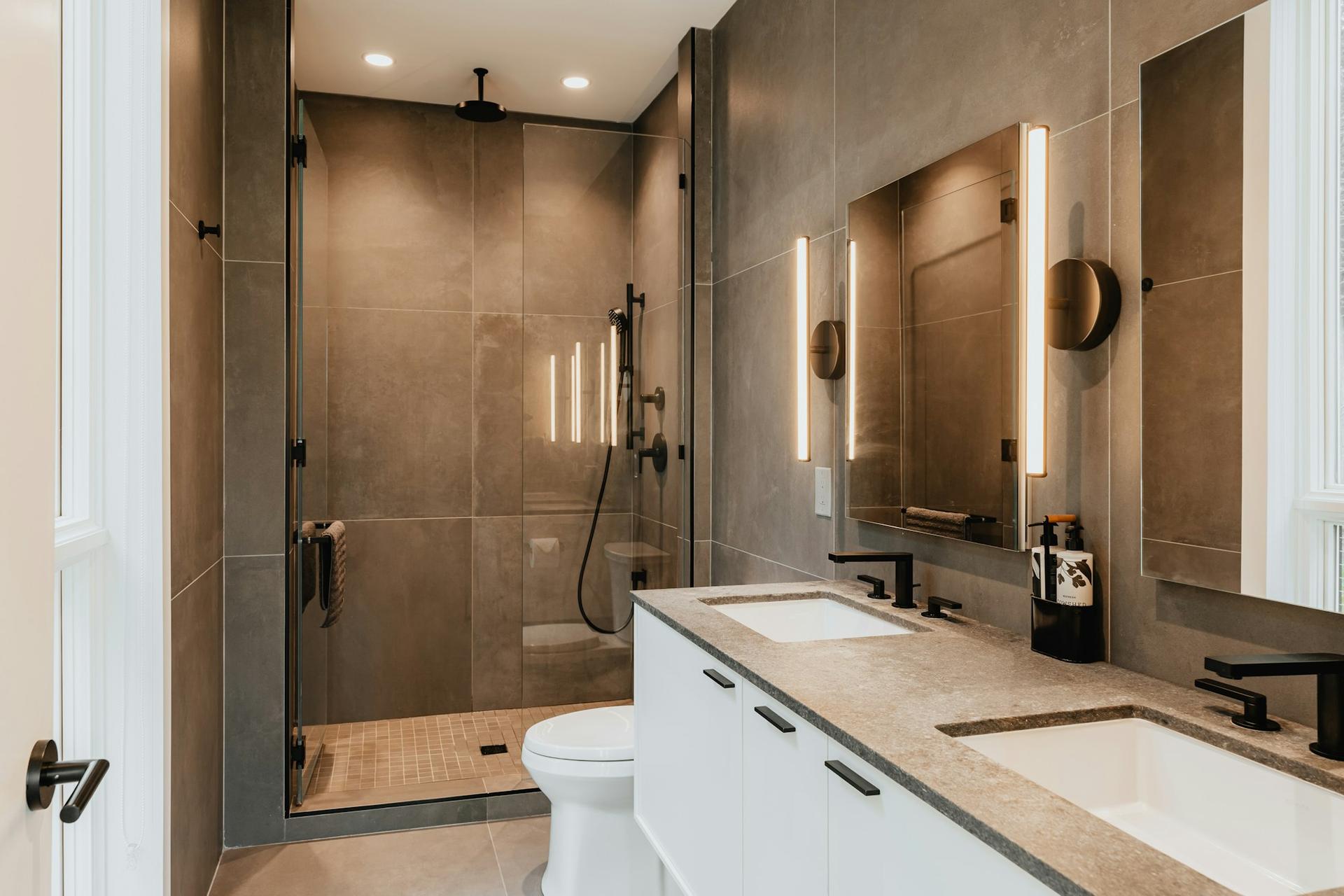 A renovated bathroom with large grey wall tiles, a glass walk-in shower and a double vanity with a stone worktop.
