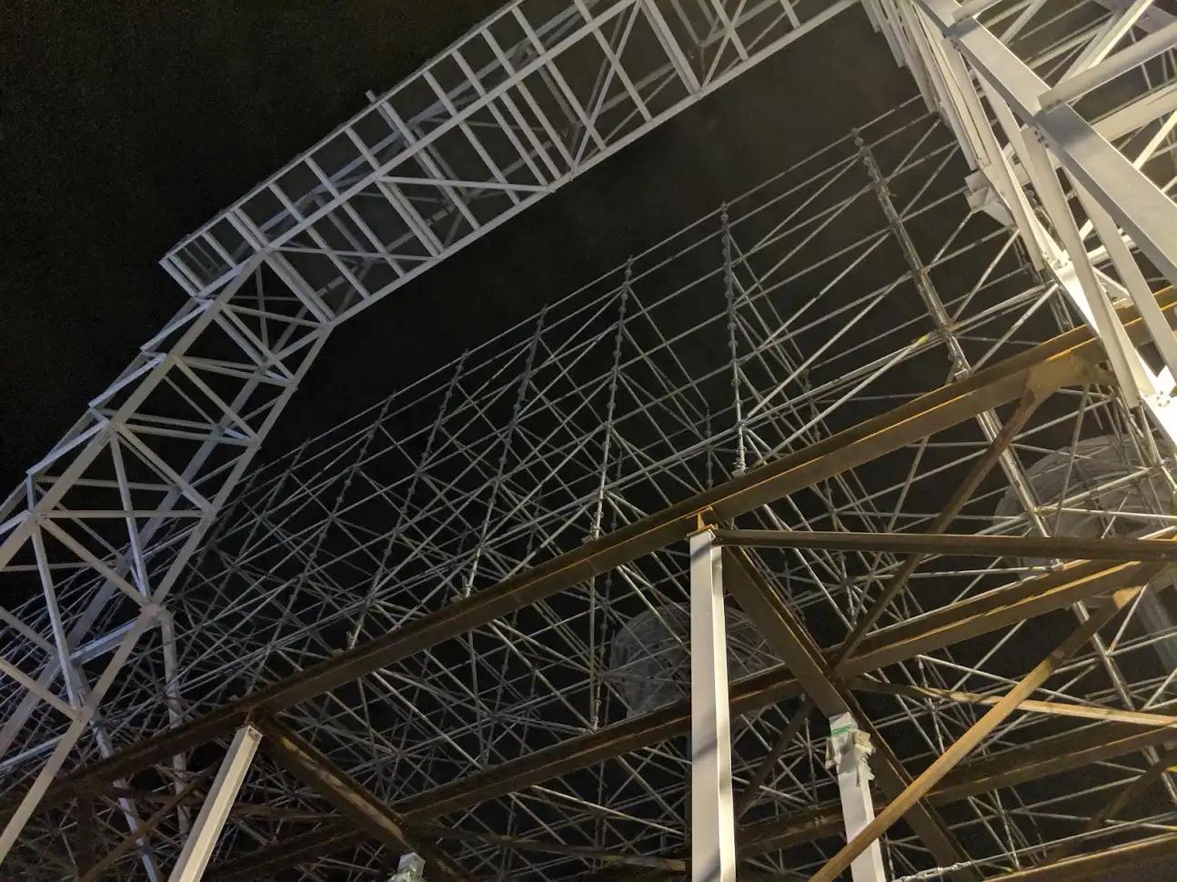 Large metal scaffolding structure photographed at night, with intersecting beams and supports forming a complex geometric framework.