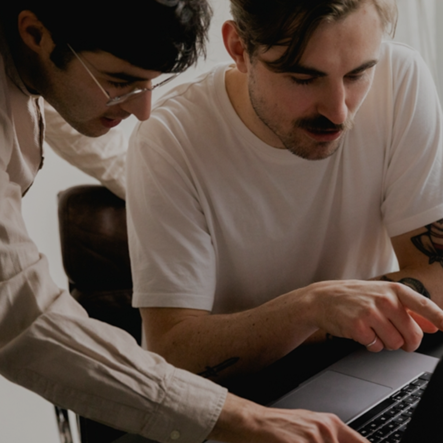 Two people collaborating while looking at a laptop screen, one seated and pointing as the other leans in to discuss or assist.