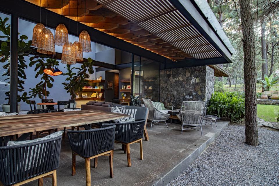 Terraza techada con mesa larga y lámparas de mimbre frente al bosque