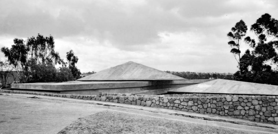 Casa de cubiertas piramidales bajas detrás de un muro de piedra, en blanco y negro