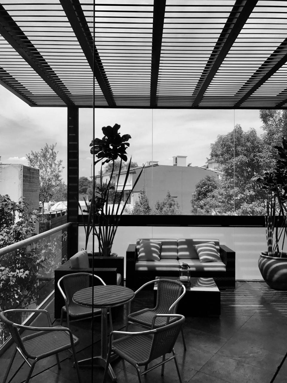 Terraza bajo pérgola de lamas con sala y comedor, en blanco y negro