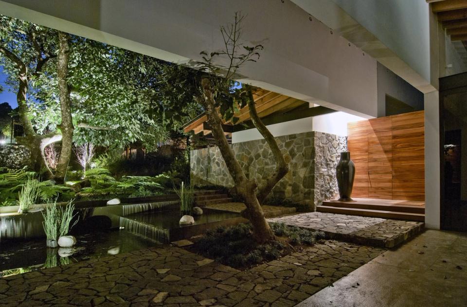 Patio iluminado de noche con espejo de agua, árbol y muro de piedra
