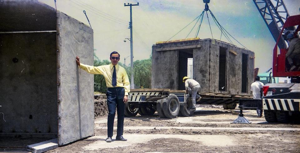 Fotografía de obra: un hombre junto a un panel prefabricado mientras una grúa eleva un módulo