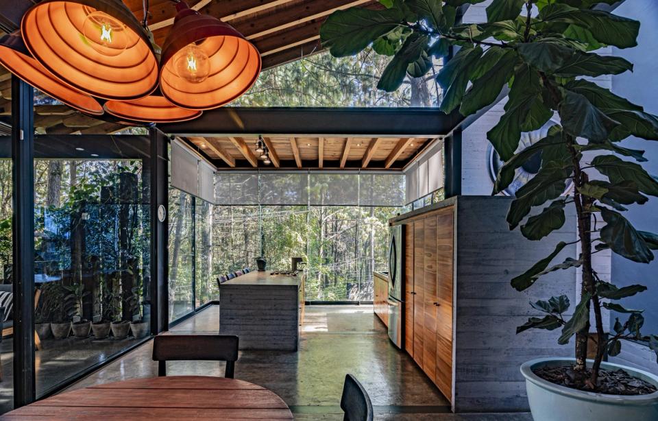 Comedor y cocina con lámparas naranjas y muros de cristal hacia el bosque
