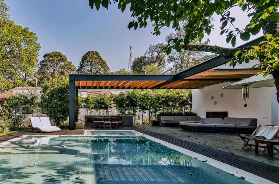 Alberca frente a la pérgola de madera y la sala exterior con chimenea